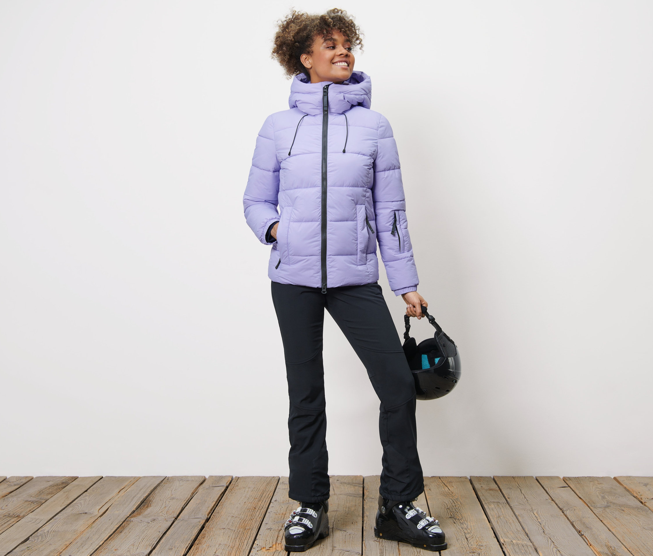 Kobieta w pikowanej kurtce narciarskiej, czarnych spodniach i butach narciarskich trzyma kask.