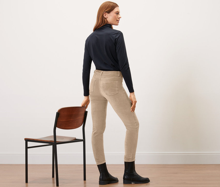 Kobieta stoi bokiem opierając się o krzesło, ma na sobie karmelowe spodnie z aksamitu.