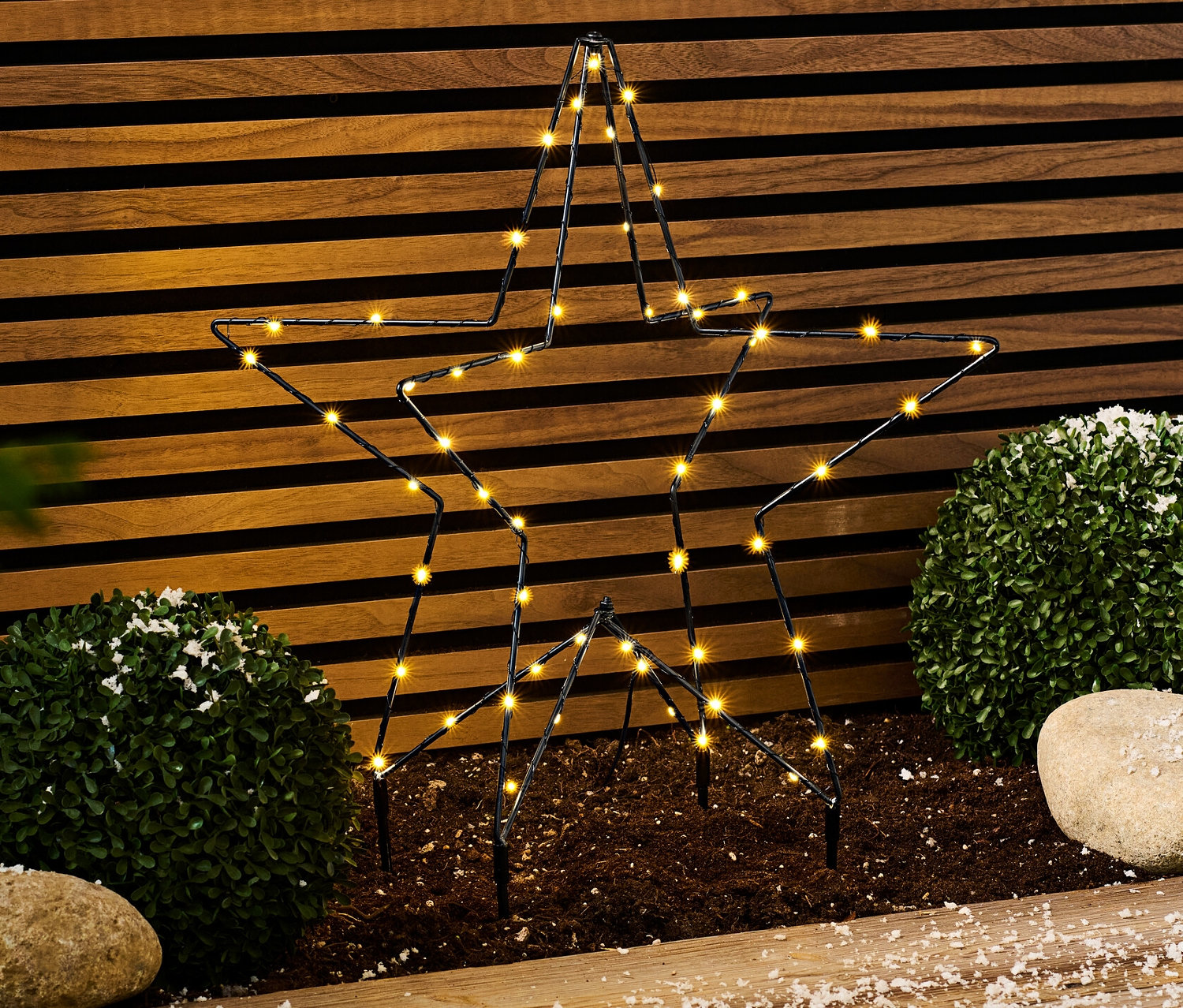 Metalowa gwiazda LED do użytku na zewnątrz stoi na ziemi obok krzewów i kamieni.