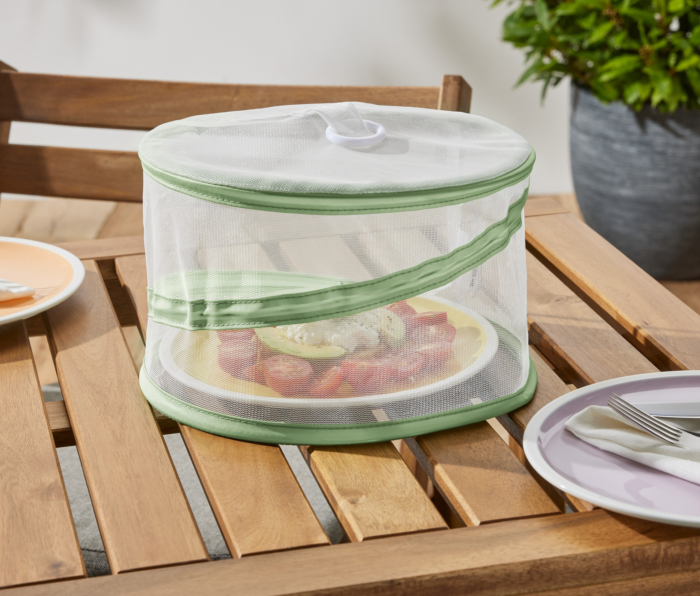 Składana osłona na żywność, mała stoi na drewnianym stole i chroni talerz z jedzeniem.