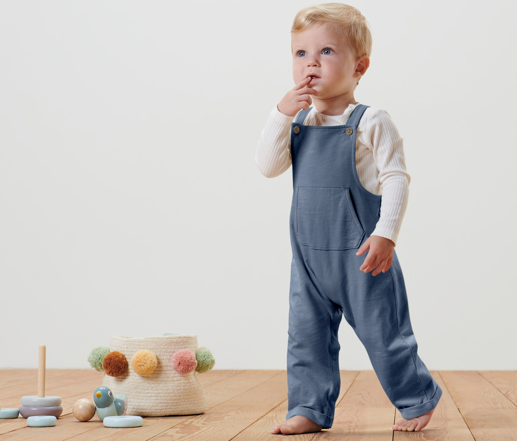 Małe dziecko stoi na drewnianej podłodze, ubrane w niebieskie ogrodniczki niemowlęce.