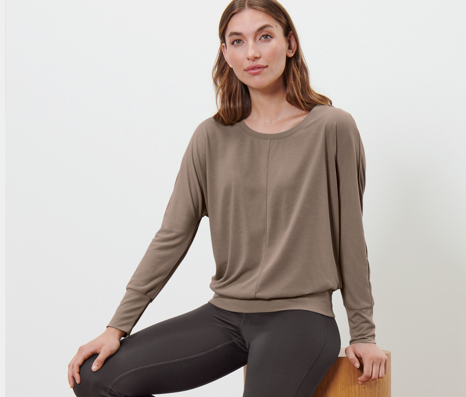 Kobieta siedzi na drewnianych stopniach, ubrana w szarobrązową koszulkę do sportu i jogi.