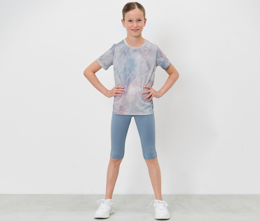 Dziewczynka w odzieży sportowej pozuje z rękami na biodrach, ma na sobie legginsy sportowe o długości 3/4 i T-shirt.