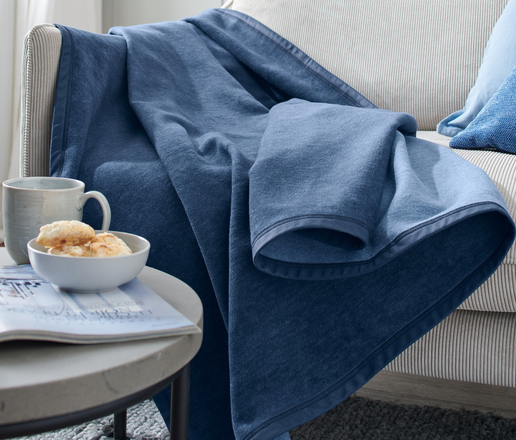 Na kanapie leży niebieski koc. Na stoliku obok kanapy stoi filiżanka, miska z jedzeniem i czasopismo.