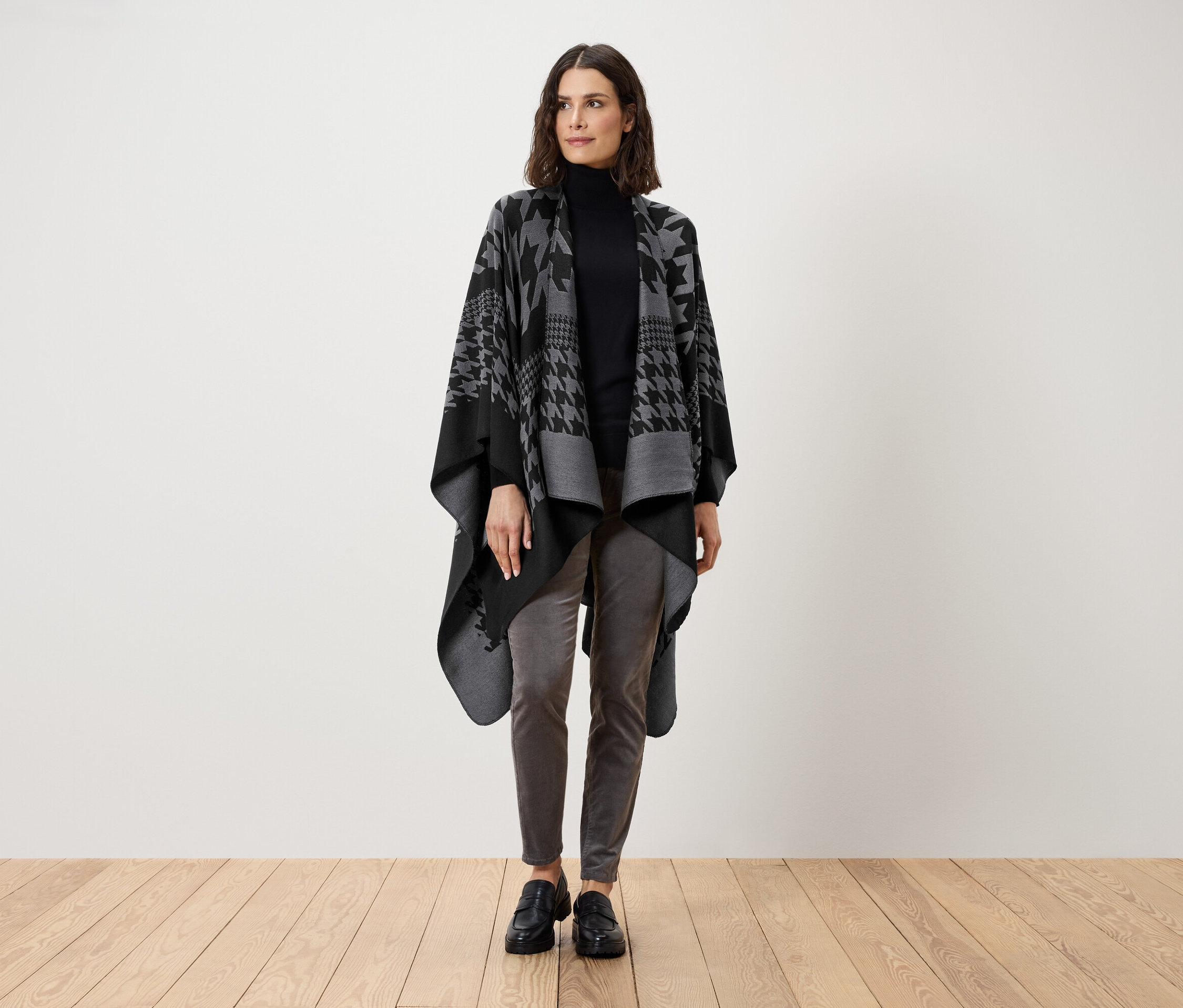Kobieta stoi w czarnym golfie, szarych spodniach i szaro-czarnej pelerynie w pepitkę.