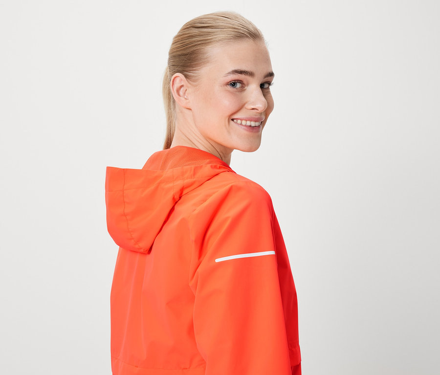 Uśmiechnięta kobieta patrzy przez ramię, ma na sobie pomarańczową kurtkę z kapturem i odblaskowym paskiem.