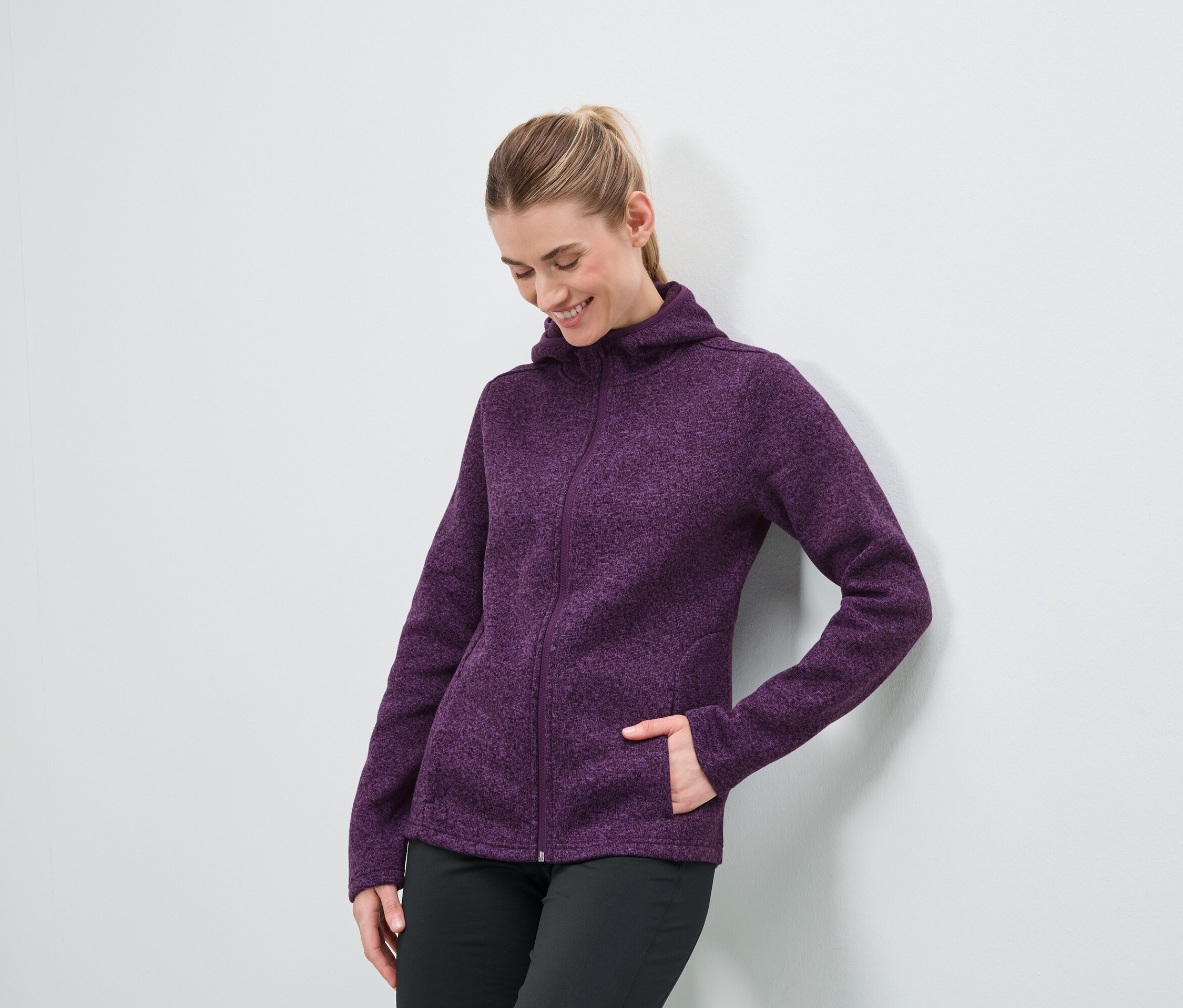 Kobieta w purpurowej bluzie z kapturem na zamek błyskawiczny i czarnych spodniach stoi przy białej ścianie.