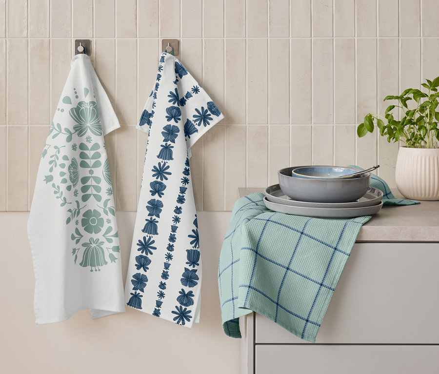 Dwie ściereczki kuchenne z kwiatowymi wzorami wiszą obok siebie na ścianie. Poniżej leży złożona zielona ściereczka z niebieskimi paskami, na której ułożone są talerze i miska. Obok stoi doniczka z ziołami.