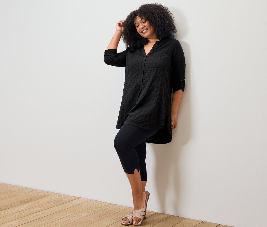 Kobieta pozuje w czarnej, długiej tunice z tkaniny i legginsach o długości 3/4 z detalami.