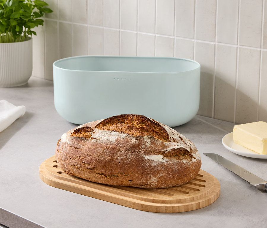 Bochenek chleba na drewnianej desce obok niebieskiego chlebaka, kawałek masła i nóż na blacie.