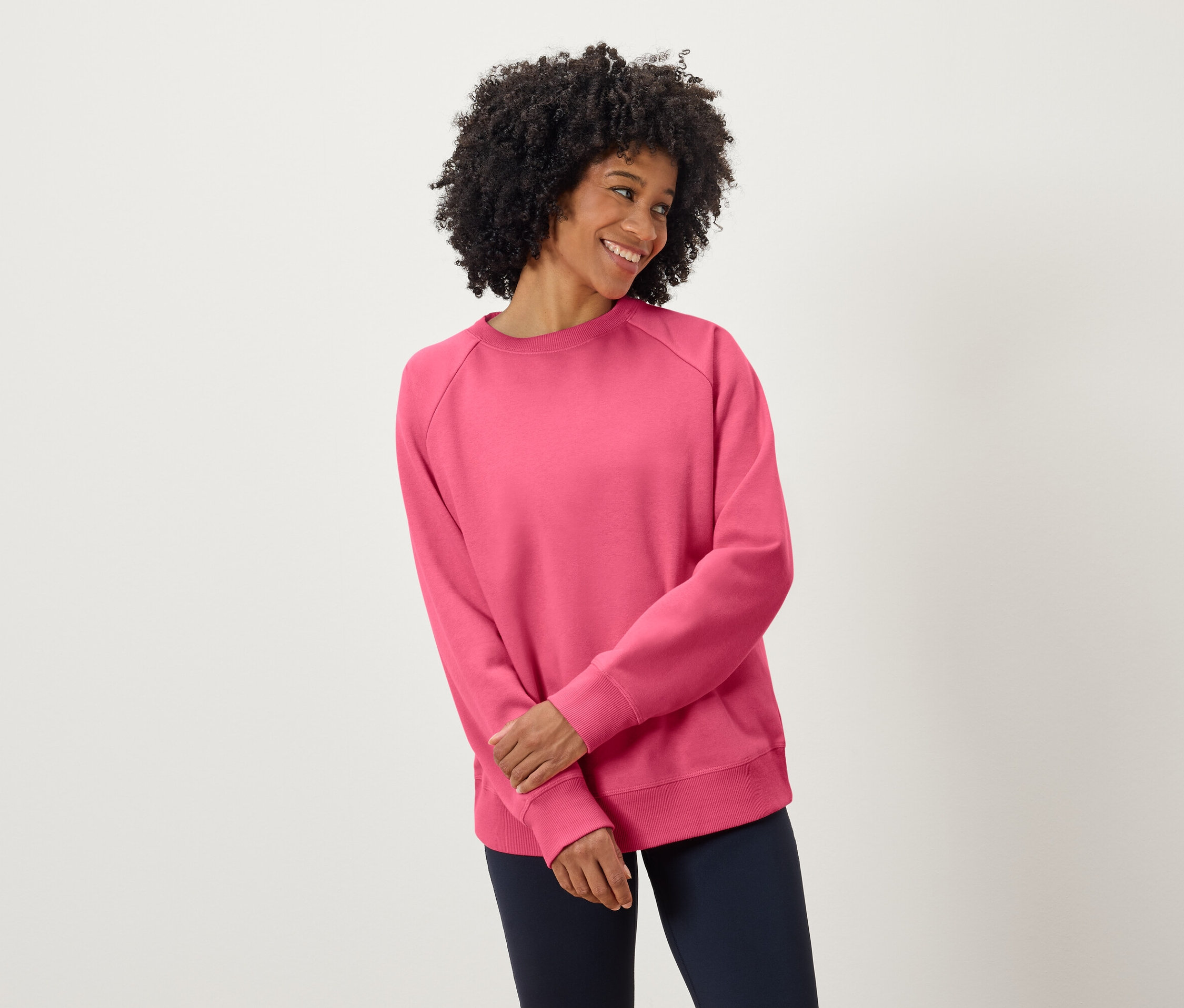 Kobieta z ciemnymi kręconymi włosami uśmiecha się, ubrana w różową bluzę i granatowe spodnie.