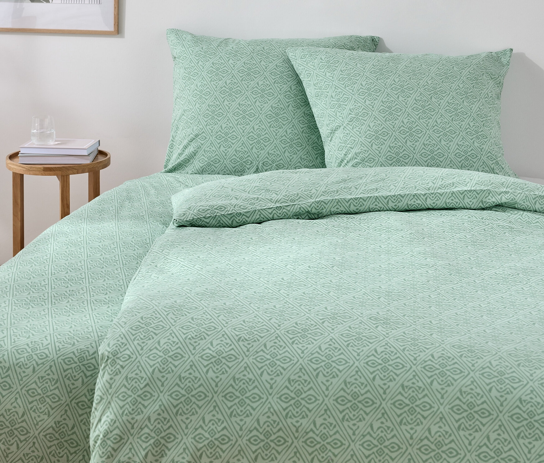 Zielona pościel jersey w standardowym i ponadwymiarowym rozmiarze na łóżku z dwiema poduszkami i drewnianym stolikiem.