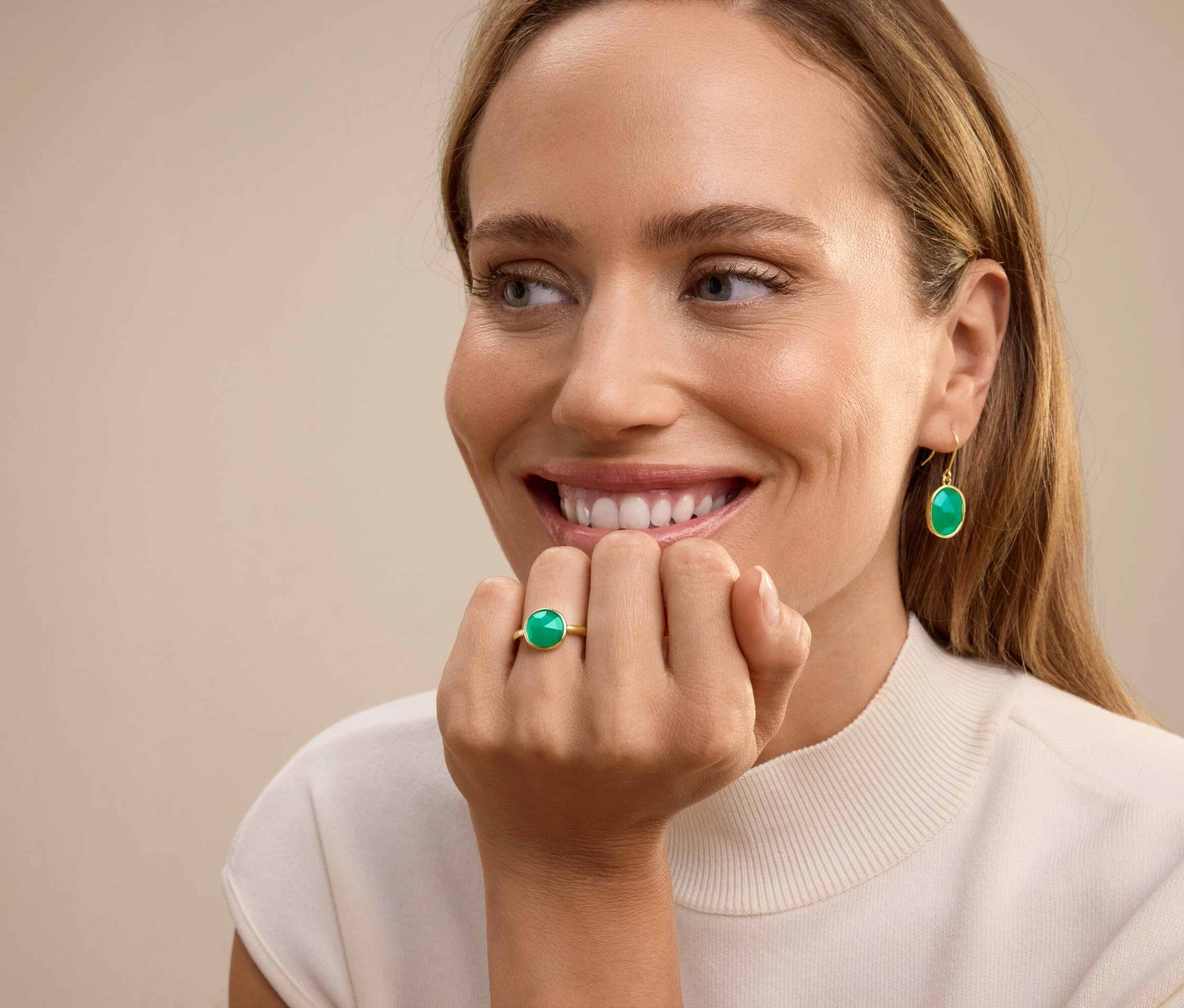 Uśmiechnięta kobieta z pierścionkiem z zielonym kamieniem na palcu serdecznym i wiszącymi kolczykami z zielonym kamieniem.