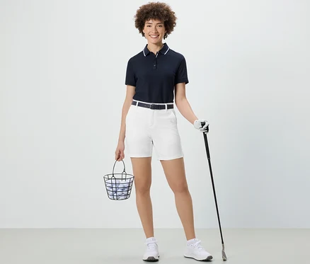 Kobieta trzyma koszyk z piłkami golfowymi i kijem golfowym. Ma na sobie białe szorty funkcyjne z softshellu.