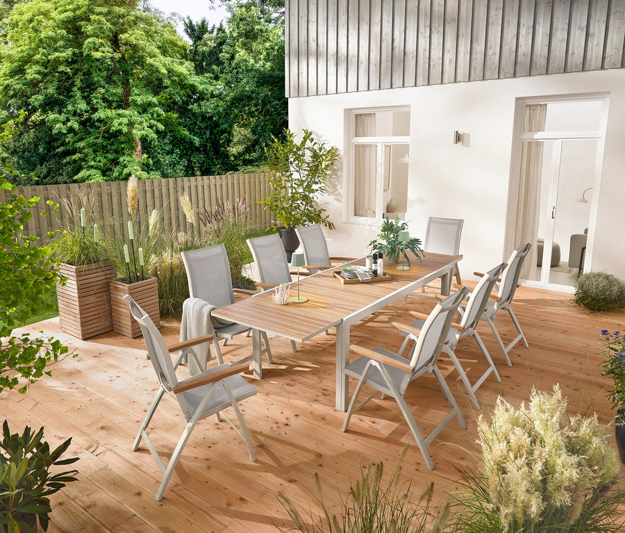 Rozkładany stół ogrodowy z aluminium »Liska« o długości ok. 200–300 cm stoi na drewnianym tarasie, otoczony sześcioma wysokimi krzesłami »Liska« z regulowanym oparciem. Widoczne są również 2 donice-kolumny na rośliny »Tinus« i 5 ogrodowych pochodni LED.