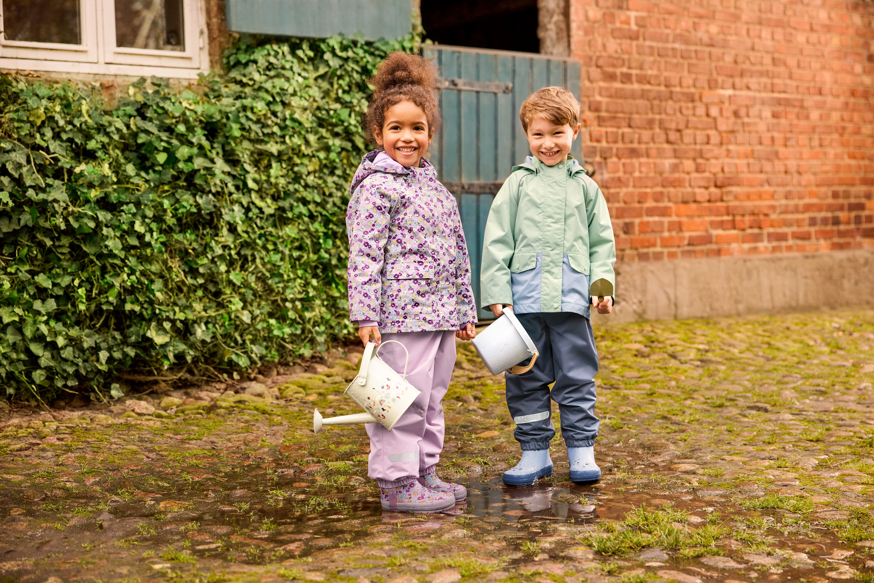 Dwoje dzieci stoi na zewnątrz: dziewczynka w kwiecistej kurtce przeciwdeszczowej i kwiecistych kaloszach oraz chłopiec w zielonej kurtce przeciwdeszczowej i niebieskich kaloszach. Oboje noszą spodnie przeciwdeszczowe.