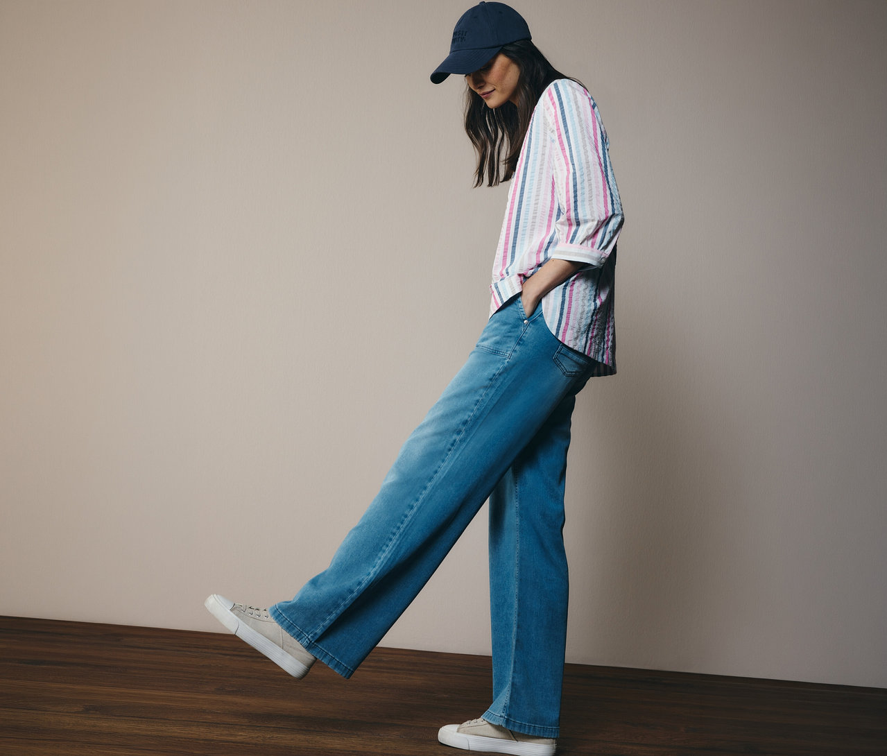 Kobieta w dżinsach i czapce bejsbolówce ma na sobie bluzkę koszulową CECIL w paski. Stoi na drewnianej podłodze.