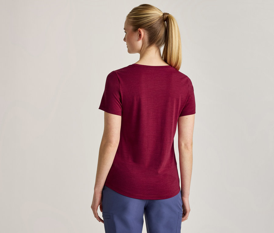 Kobieta widziana od tyłu, ubrana w bordowy T-shirt z krótkim rękawem i niebieskie spodnie. Jej blond włosy są związane w kucyk.