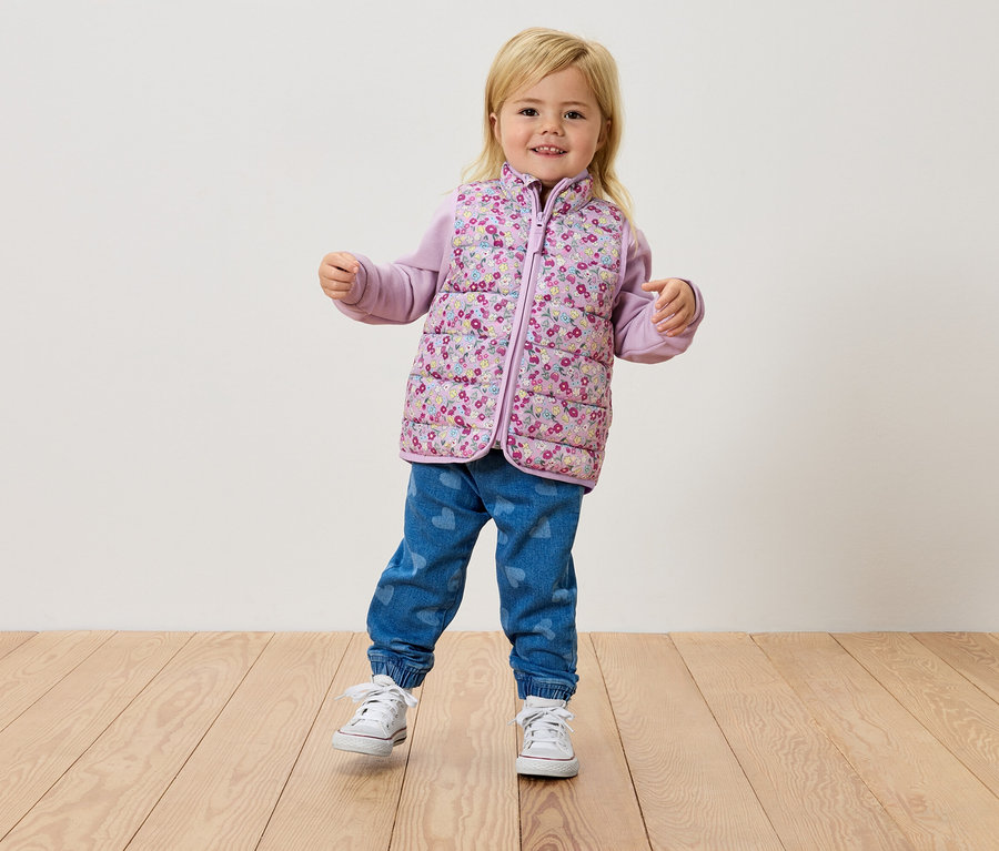 Mała blond dziewczynka stoi na drewnianej podłodze, ubrana w kwiecistą kamizelkę, fioletową bluzkę i dżinsy.