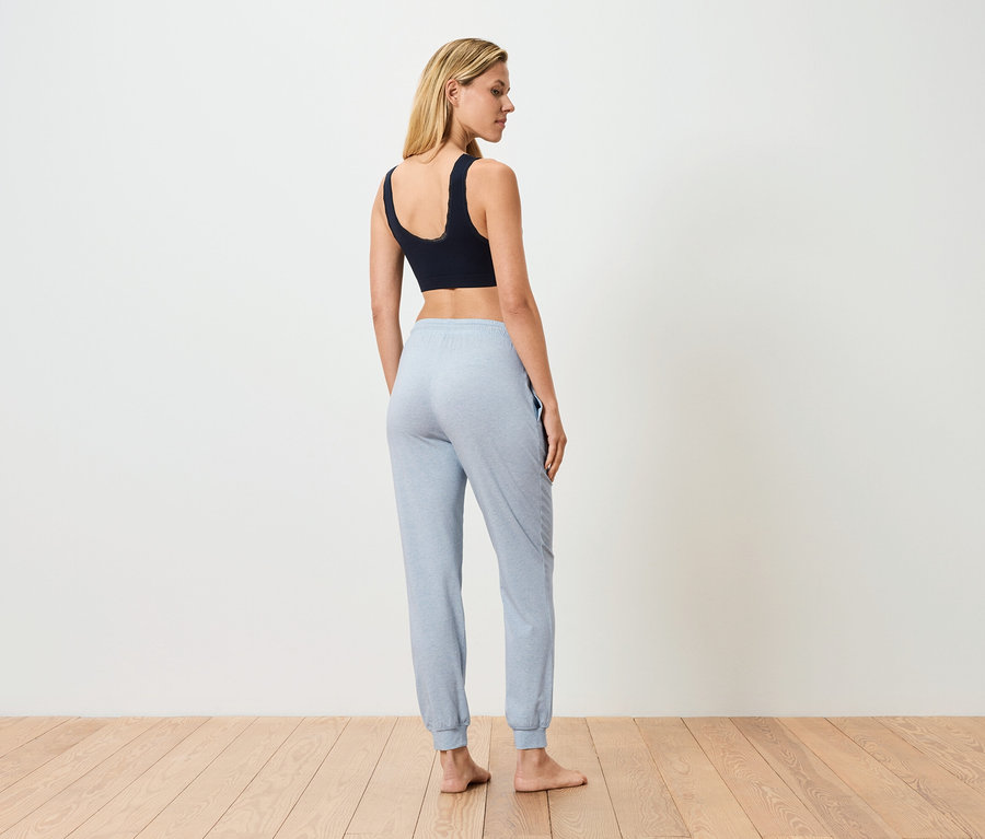 Kobieta stoi tyłem, ubrana w czarny biustonosz sportowy i jasnoniebieskie spodnie dresowe.