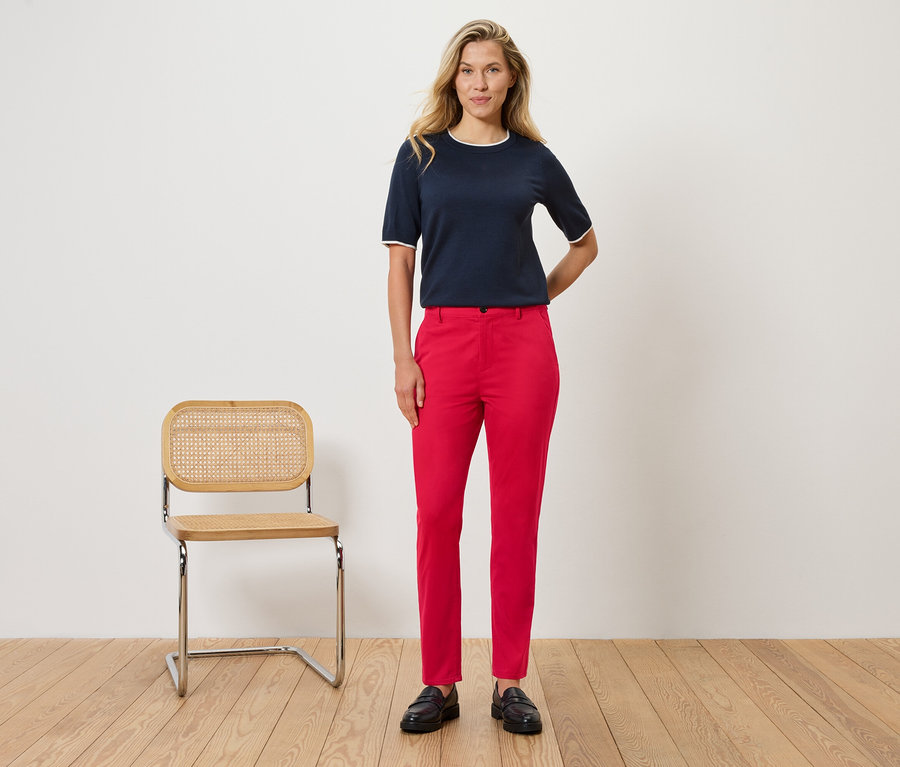 Kobieta stoi przed ścianą obok krzesła. Ma na sobie granatową bluzkę z białymi detalami i czerwone spodnie.
