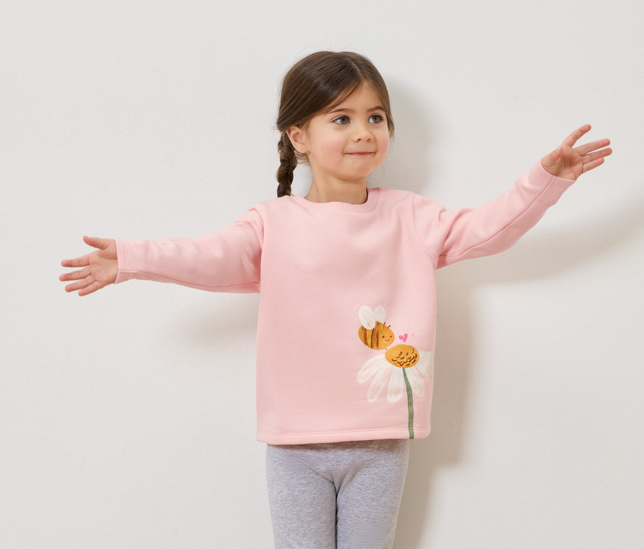 Dziewczynka z warkoczami w różowym swetrze z nadrukiem pszczoły i stokrotki, szare legginsy, z rozłożonymi ramionami.