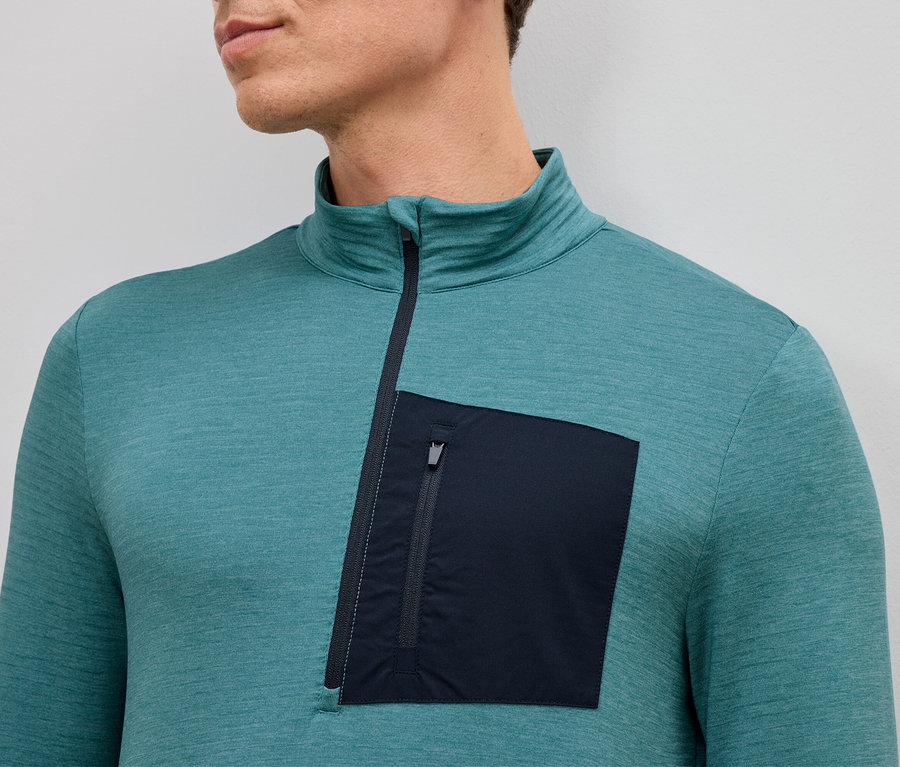 Zbliżenie mężczyzny w turkusowej koszuli z długim rękawem, krótkim zamkiem błyskawicznym i pojedynczą naszytą kieszenią zapinaną na zamek po lewej stronie.