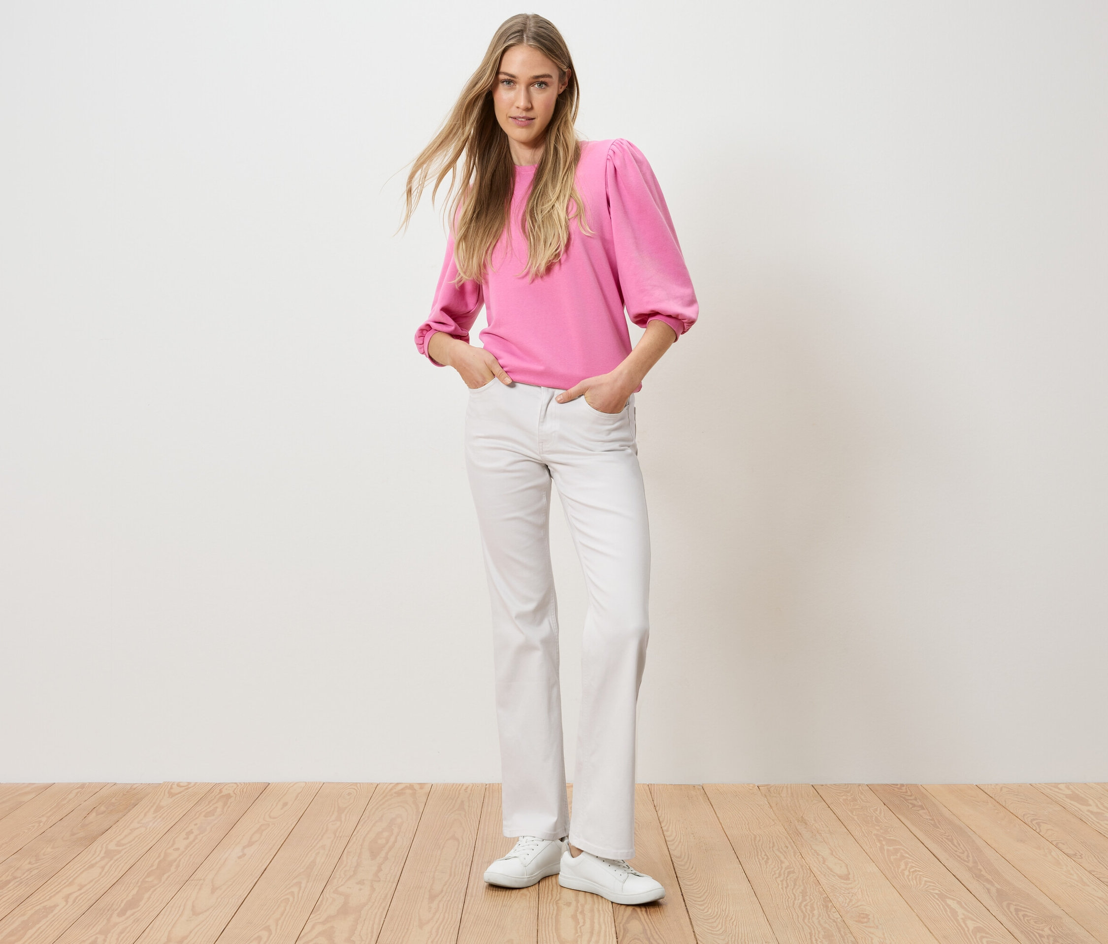 Kobieta stoi z rękami w kieszeniach spodni. Ma na sobie różowy top i białe dżinsy.