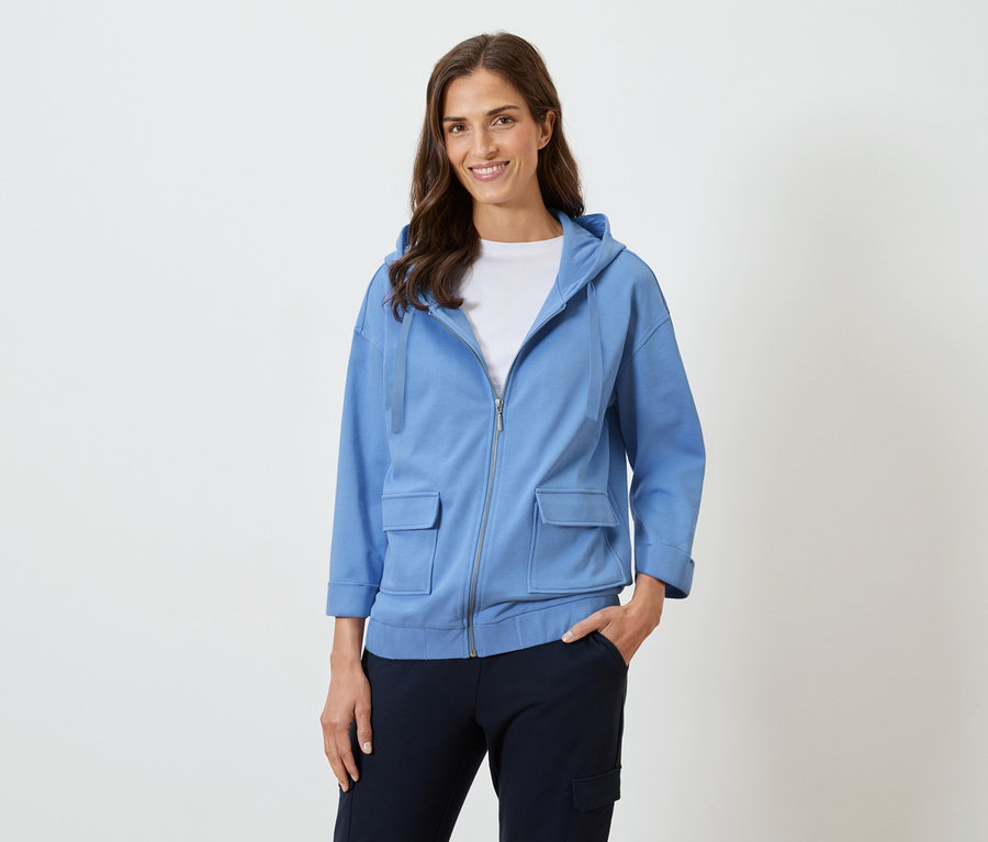 Uśmiechnięta kobieta z długimi brązowymi włosami w niebieskiej bluzie z kapturem i ciemnych spodniach.