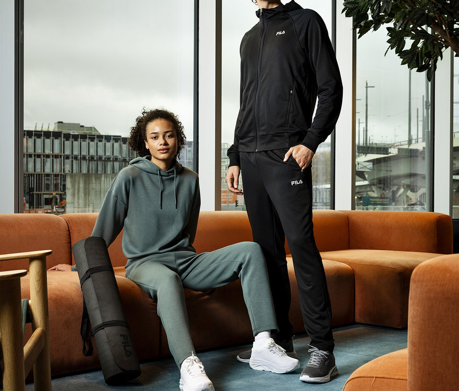 Młoda kobieta siedzi z zwiniętą matą do jogi obok siebie na pomarańczowej sofie, obok stoi młody mężczyzna. Oboje mają na sobie dresy i trampki.