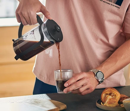 Mężczyzna nalewa kawę z czarnego zaparzacza tłokowego 300 ml do szklanki. Na stole leży croissant.