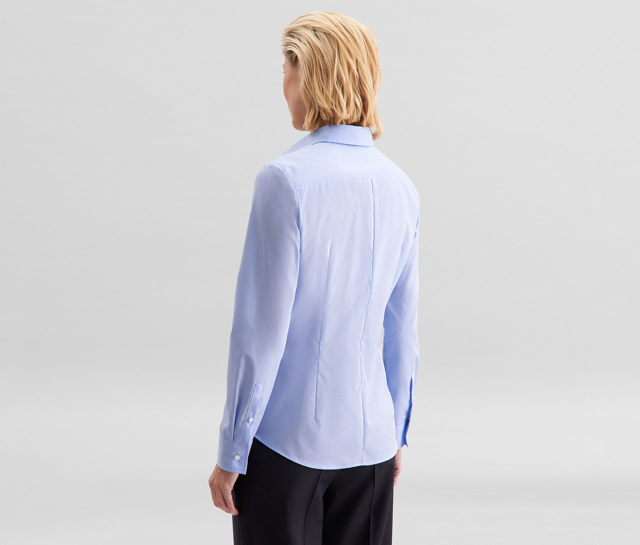 Kobieta od tyłu, z krótkimi blond włosami, ma na sobie jasnoniebieską bluzkę Seidensticker Slim Fit non iron i czarne spodnie.