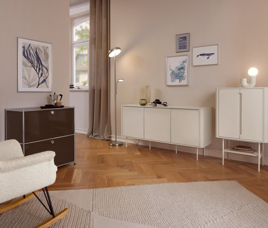 Przytulny salon z jasnymi ścianami i drewnianą podłogą. Na pierwszym planie kremowy fotel bujany stoi obok ciemnobrązowej szafki z kawiarką. Lampa podłogowa znajduje się między długą białą komodą a wyższą białą szafką. Różne oprawione dzieła sztuki zdobią ściany, a jasny dywan pokrywa część podłogi.