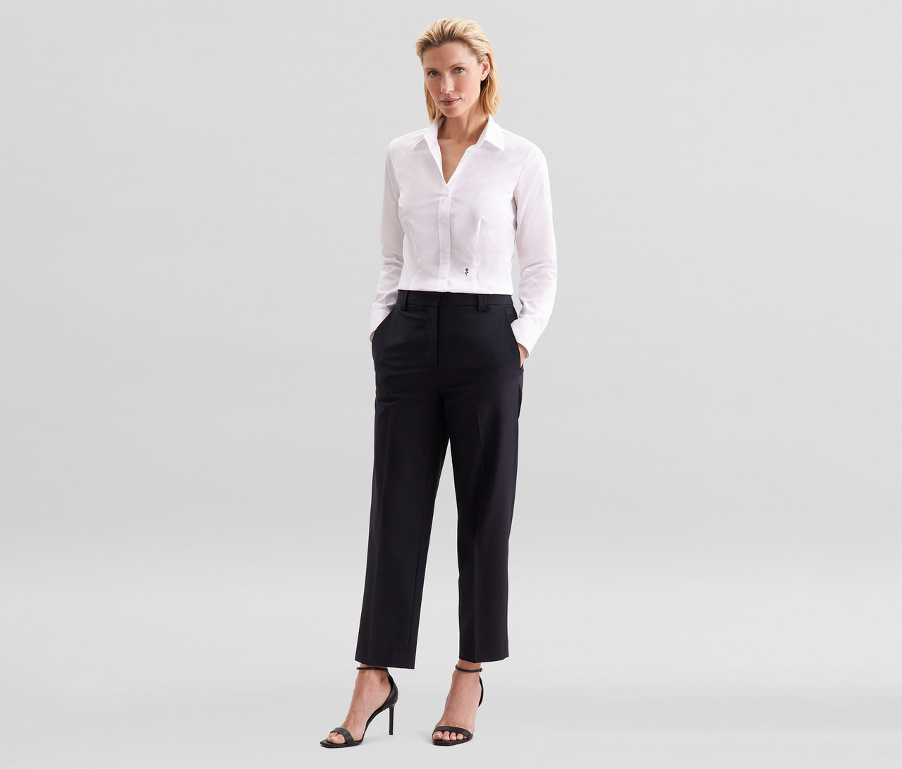 Kobieta z blond włosami stoi z rękami w kieszeniach czarnych spodni. Ma na sobie białą bluzkę Seidensticker Slim Fit non iron i czarne sandały na wysokim obcasie.