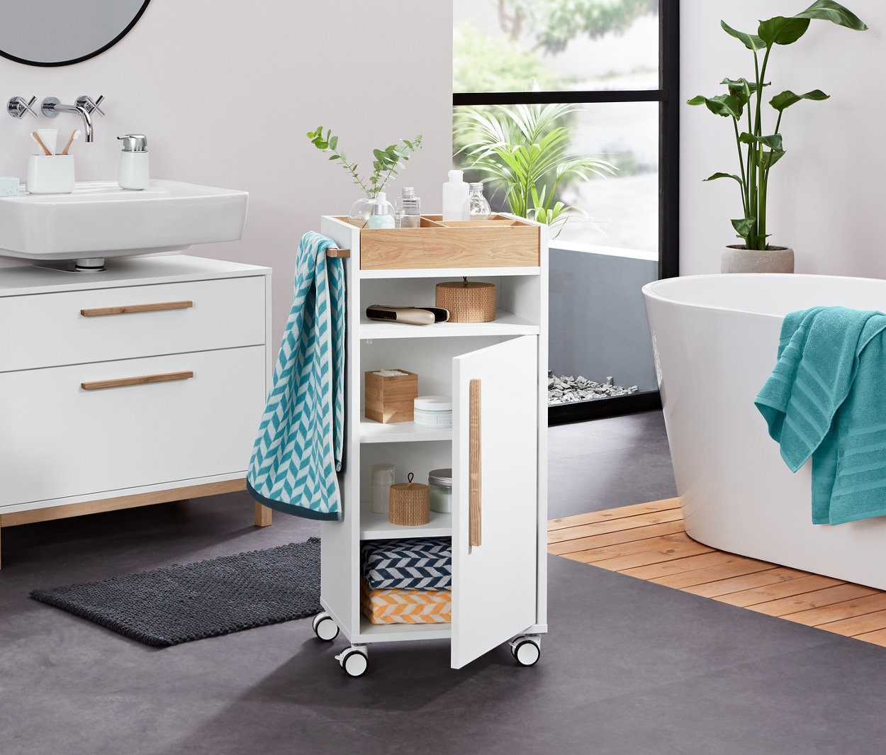 W łazience znajdują się: biała szafka pod umywalkę »Eklund« z 2 szufladami, przybornik łazienkowy, dozownik mydła, ręcznik kąpielowy frotte, ręczniki żakardowe (2 sztuki), ręczniki frotte (antracytowe, 2 sztuki), pojemniki bambusowe (2 sztuki), kamienie dekoracyjne LED (2 sztuki) i szafka łazienkowa na kółkach „Eklund”.