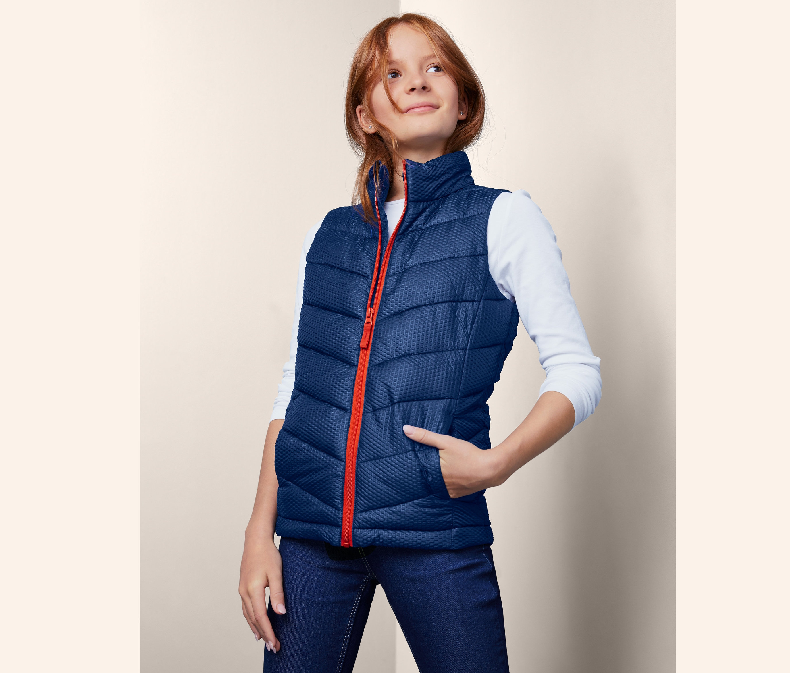 Dziewczyna ma na sobie kamizelkę pikowaną z materiału z recyklingu z pomarańczowym zamkiem i rękami w kieszeni.