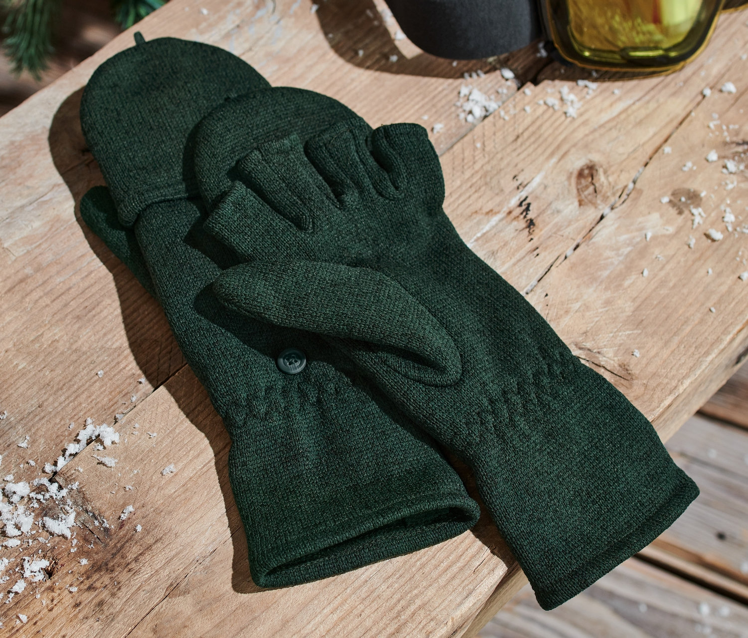 Zielone rękawice-łapawice z dzianiny polarowej leżą na drewnianej ławce obok gogli narciarskich.