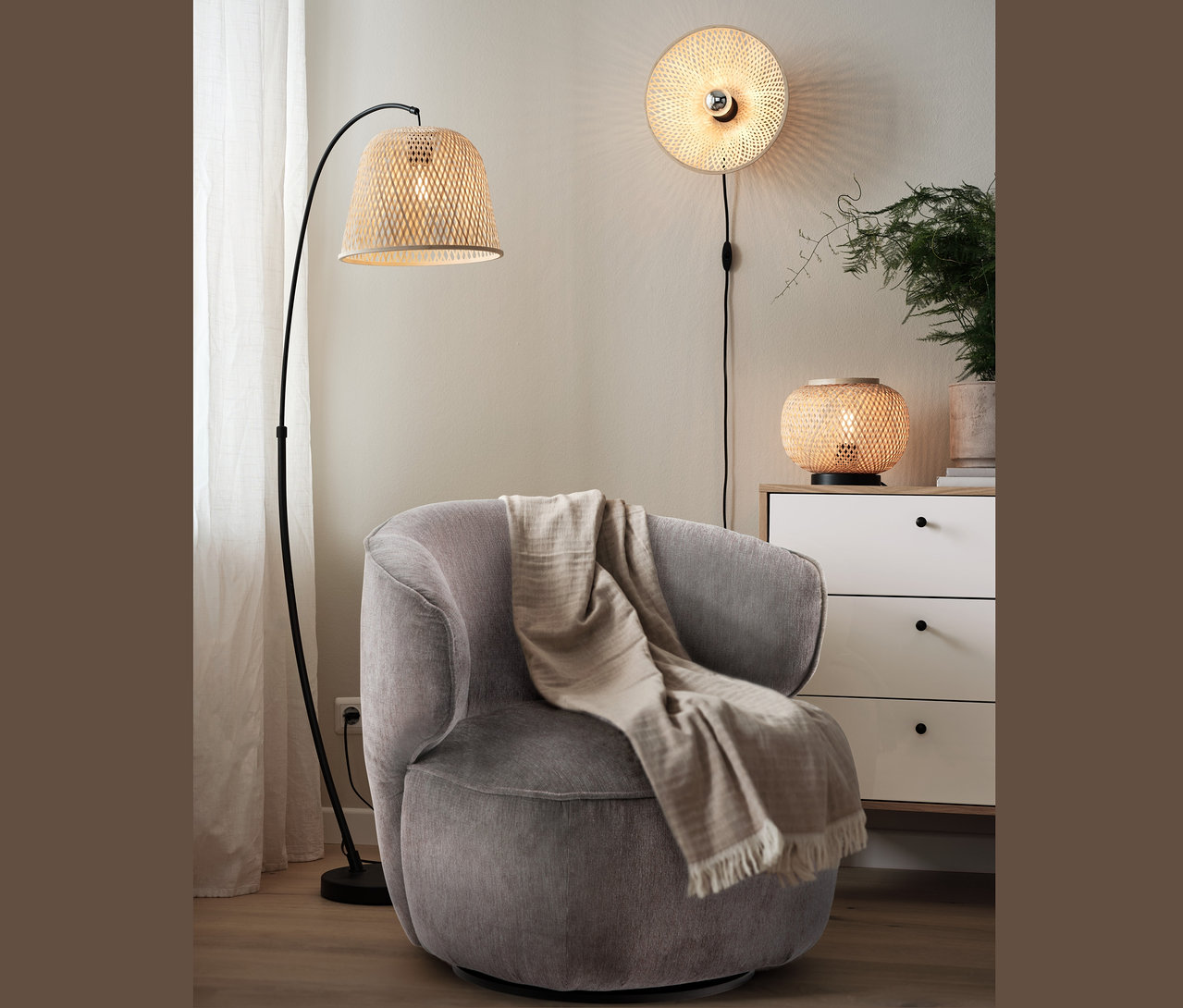 Szary fotel obrotowy z kocem stoi obok lampy stojącej z bambusowym kloszem. Na ścianie kinkiet z bambusowym kloszem, a na komodzie lampa stołowa z kloszem.