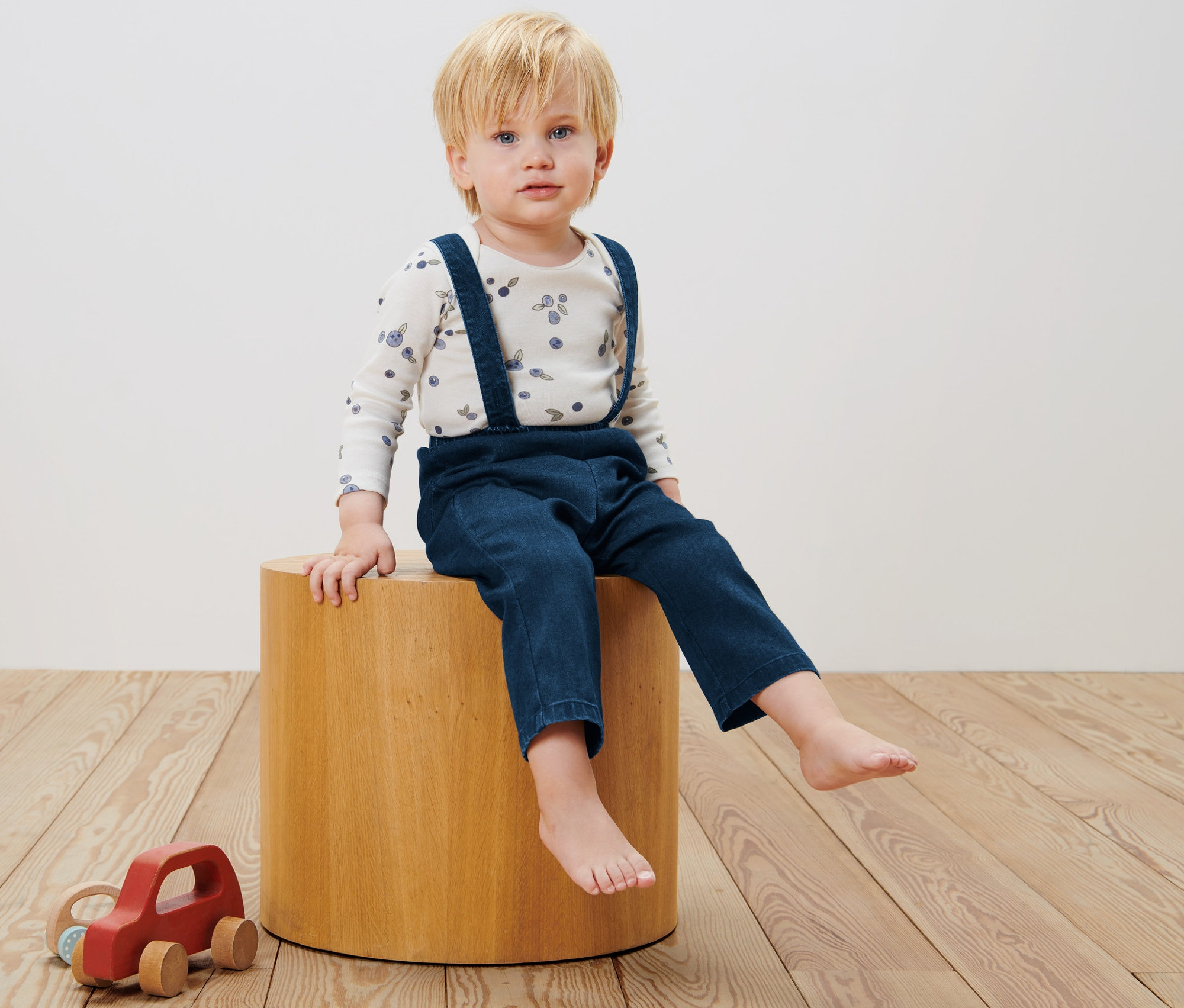 Chłopiec siedzi na drewnianym stołku i ma na sobie spodnie z szelkami.