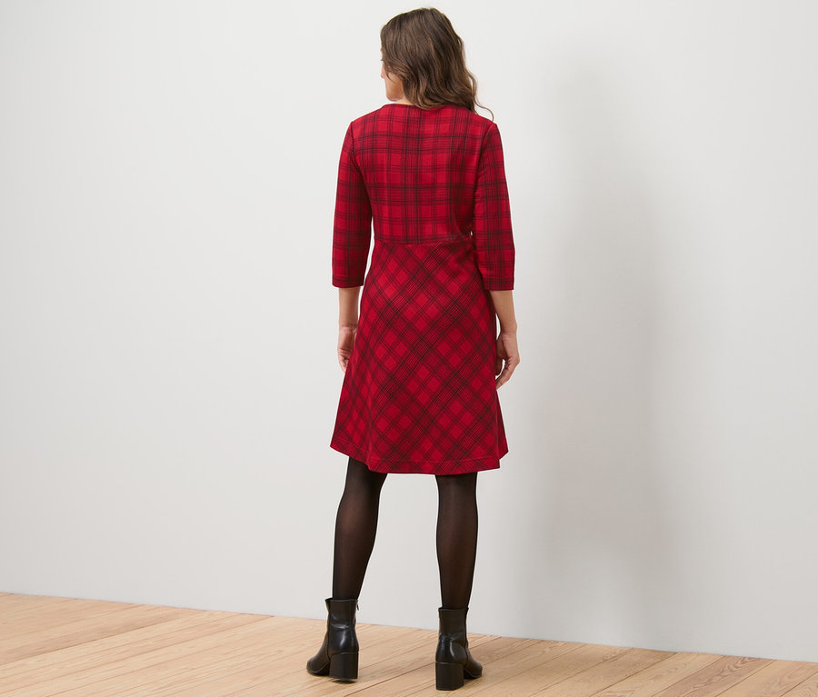 Kobieta stoi tyłem do kamery, ubrana w żakardową sukienkę w kratę, czarne rajstopy i czarne buty.