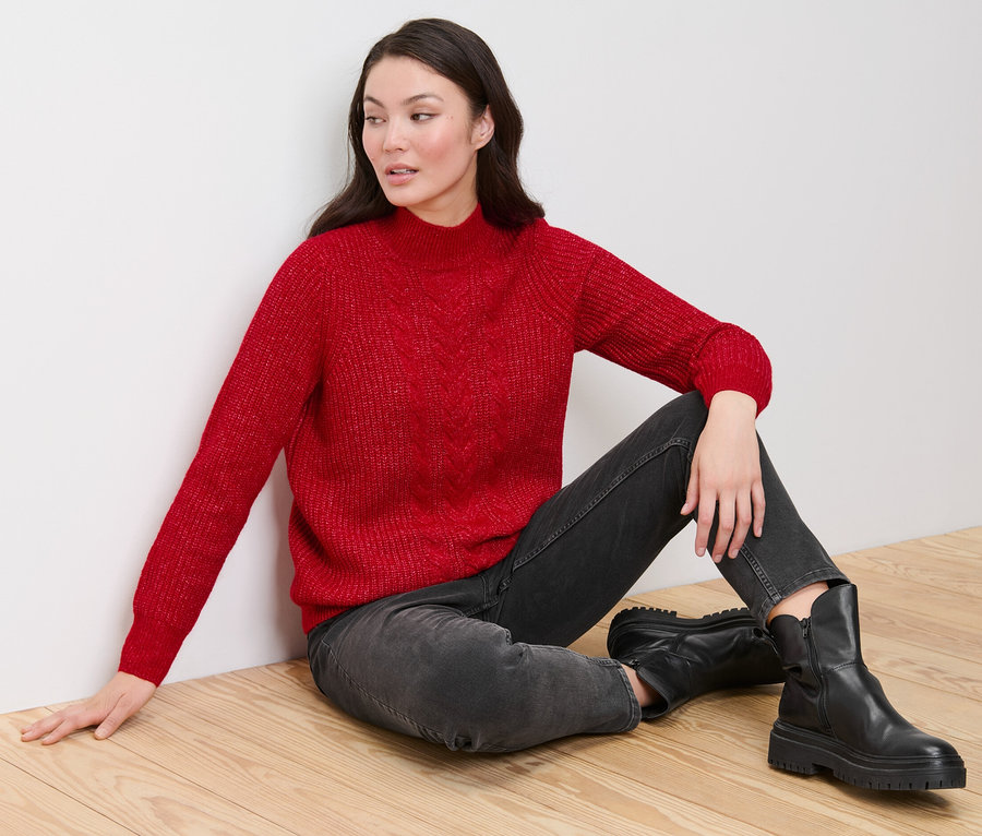 Kobieta siedzi na drewnianej podłodze i ma na sobie czerwony sweter o grubym splocie z warkoczowym wzorem, czarne dżinsy i czarne buty.