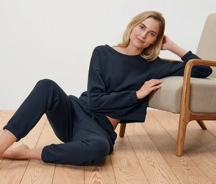 Blondynka siedzi na podłodze obok fotela, ubrana w granatowy zestaw odzieży domowej.