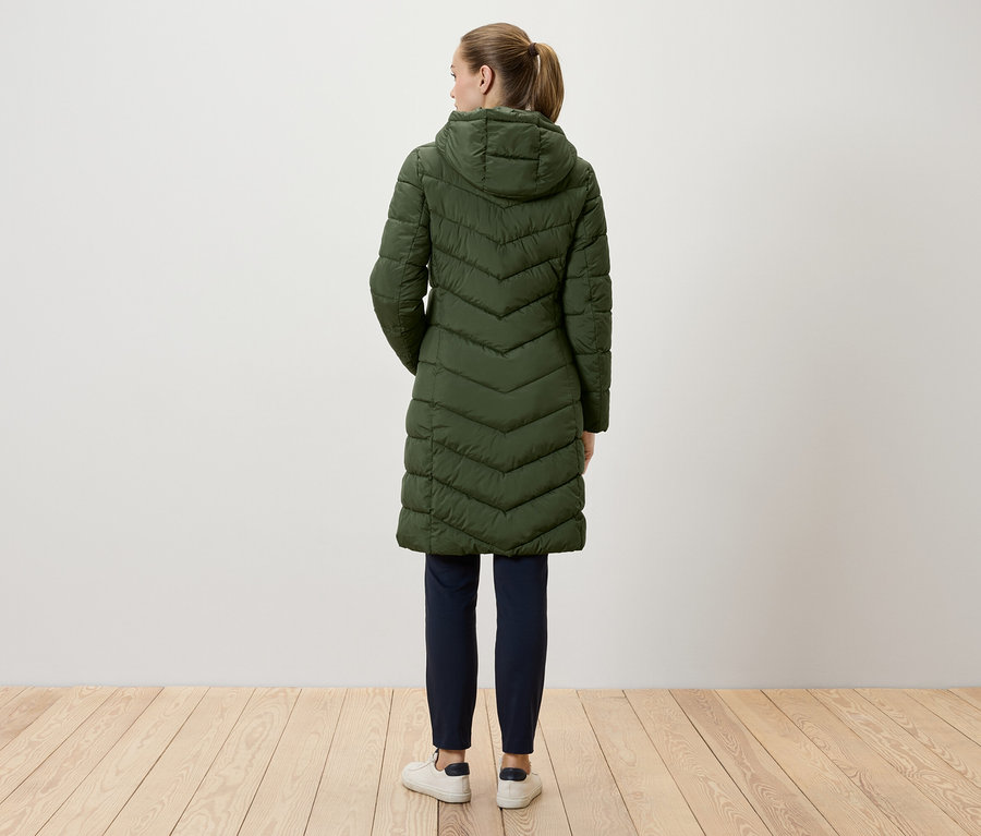 Kobieta stoi tyłem do kamery, ubrana w długi, zielony, pikowany płaszcz z kapturem, granatowe spodnie i białe tenisówki.