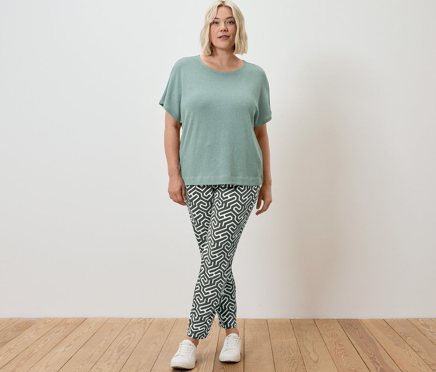 Kobieta stoi w szałwiowej koszulce z fakturowaną powierzchnią i ciemnozielonych wzorzystych spodniach z dżerseju.