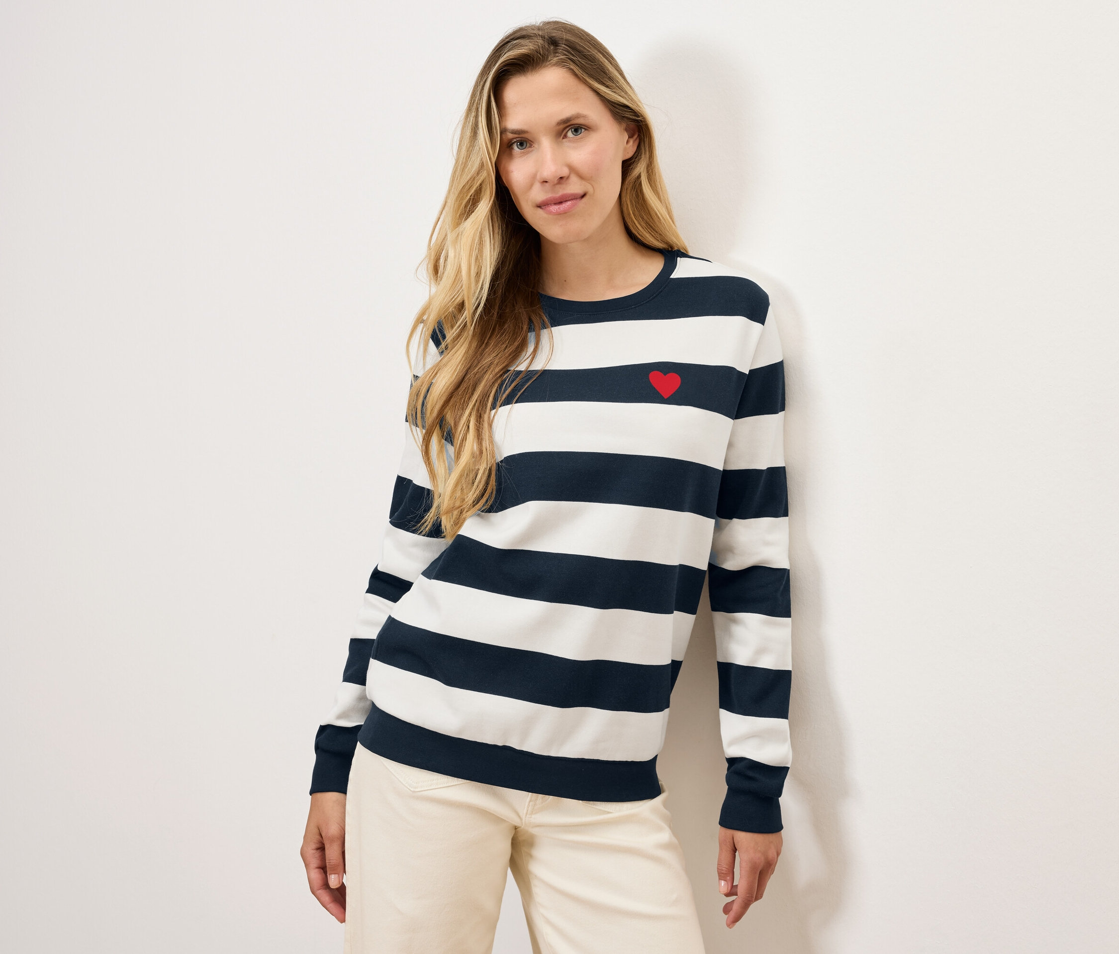 Blondynka pozuje w pasiastym swetrze z czerwonym sercem i jasnych spodniach.