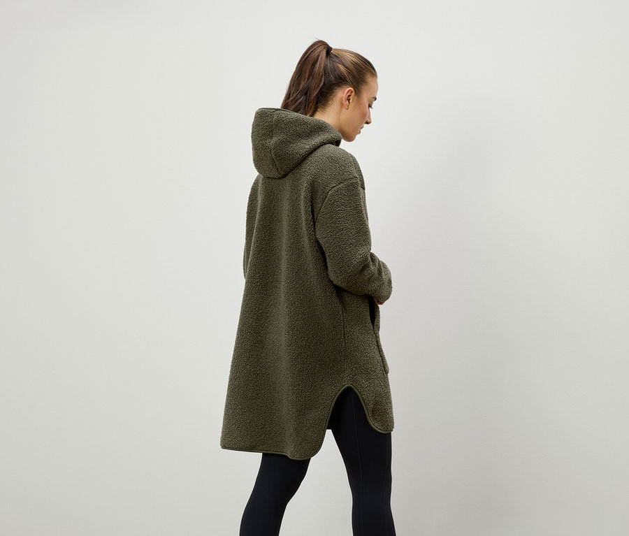 Kobieta widziana od tyłu ma na sobie oliwkowozielony, puszysty płaszcz z kapturem i czarne legginsy.