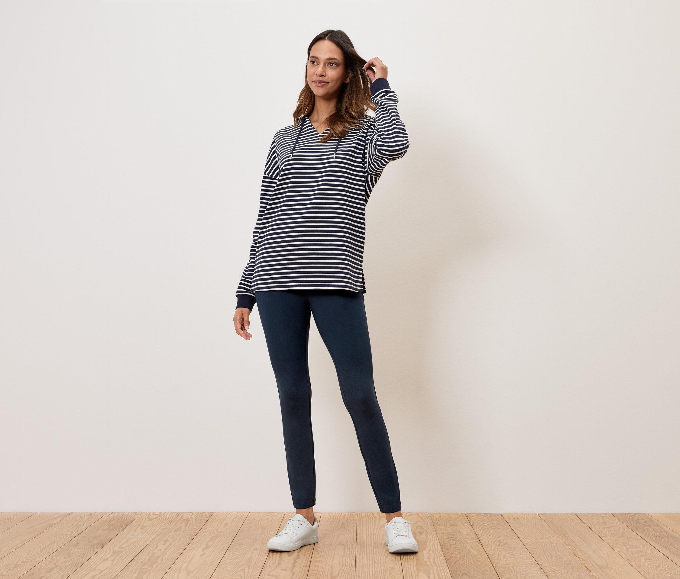 Kobieta stoi na drewnianej podłodze, ubrana w bluzkę w paski, granatowe legginsy i białe tenisówki.