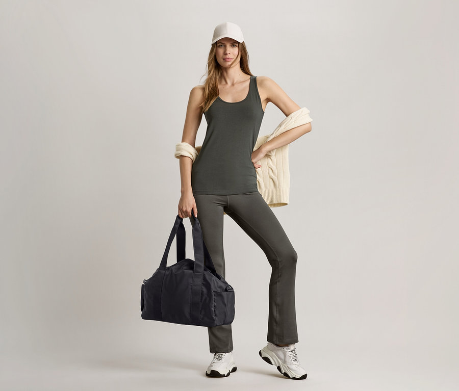 Kobieta w oliwkowym topie i spodniach sportowych modelujących sylwetkę, trzyma czarną torbę sportową.