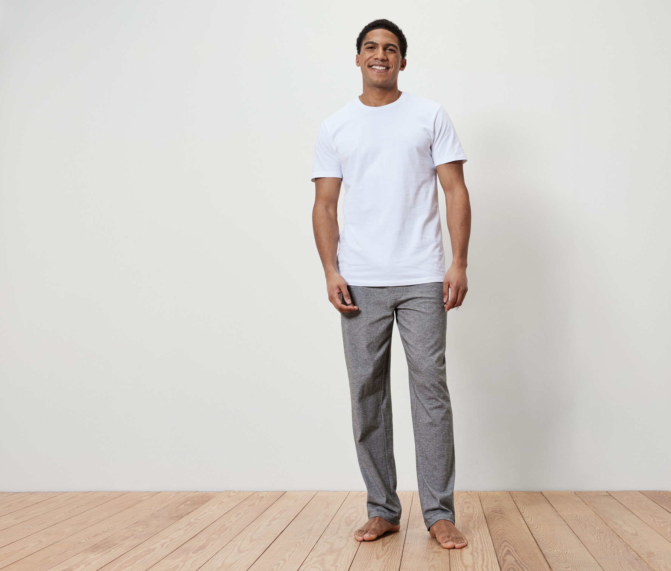 Uśmiechnięty mężczyzna stoi boso na drewnianej podłodze. Ma na sobie białą koszulkę i szare spodnie.