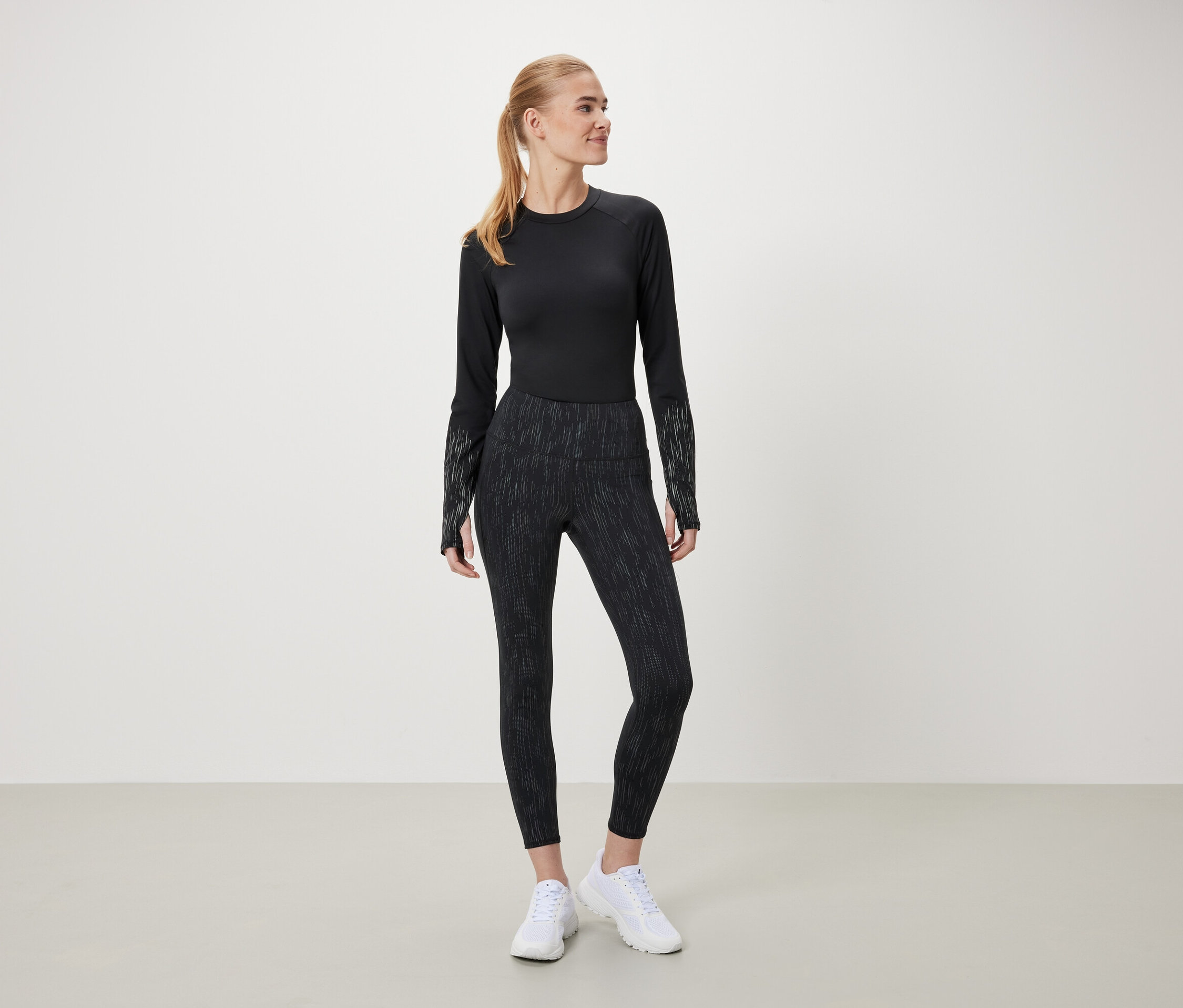 Kobieta w czarnym topie z długim rękawem i wzorzystych legginsach stoi na jasnym tle.